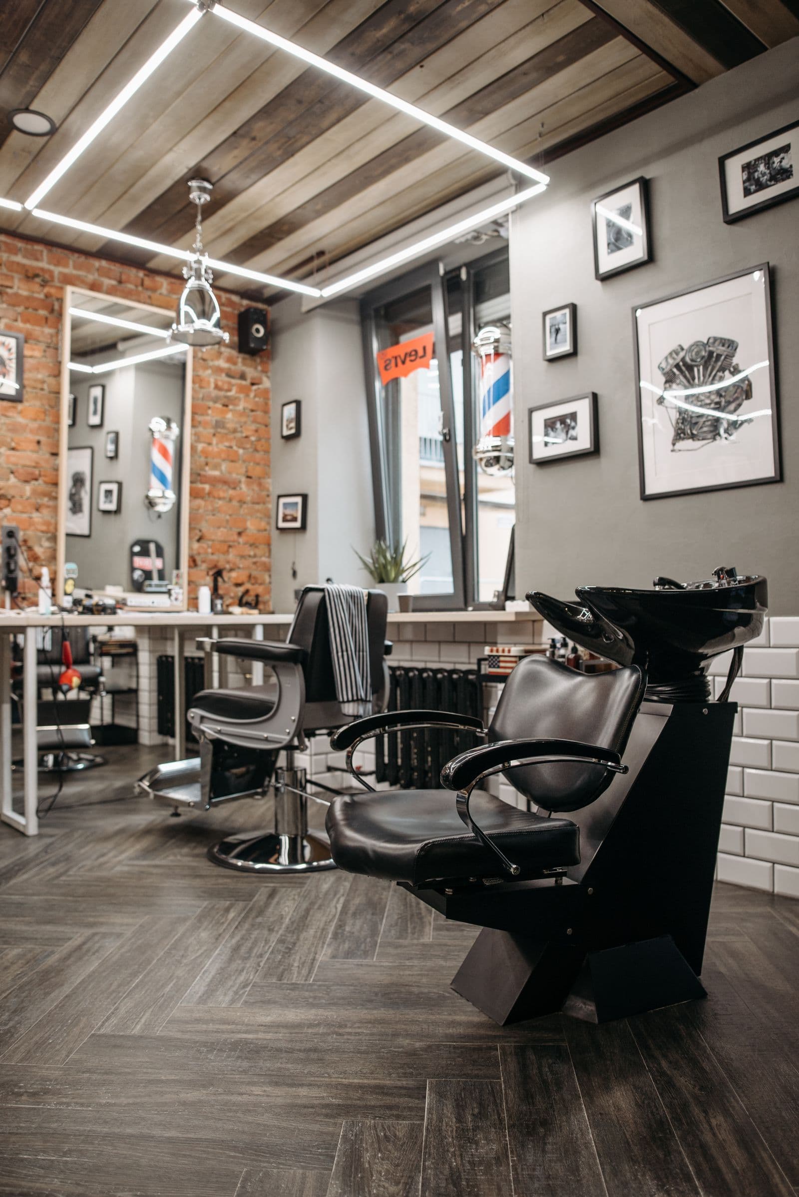 A modern barbershop interior with black leather chairs, a brick wall and warm overhead lighting, nobody in it.